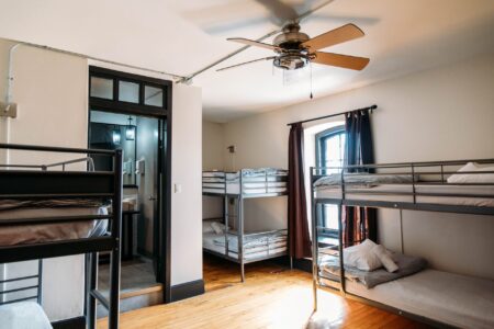 A white dorm room with 3 bunk beds with 2 beds each, two big windows and a ceiling fan